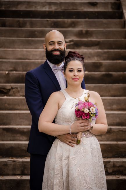A bride and groom pose together on a grand staircase with wooden steps, illuminated by soft lighting. The bride is holding a floral bouquet comprising pink roses, white spray roses, and greenery, wrap