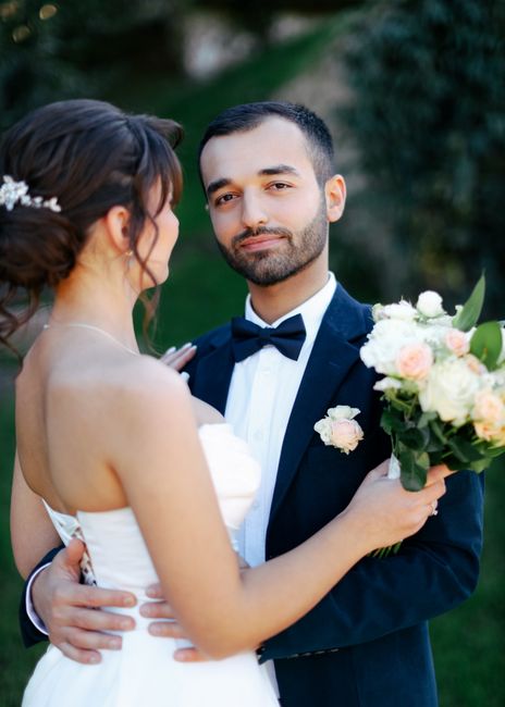 A groom in a dark suit with a white shirt and black bow tie holds a bride in a strapless white wedding dress close, both standing outdoors with lush green foliage in the background. The bride has dark