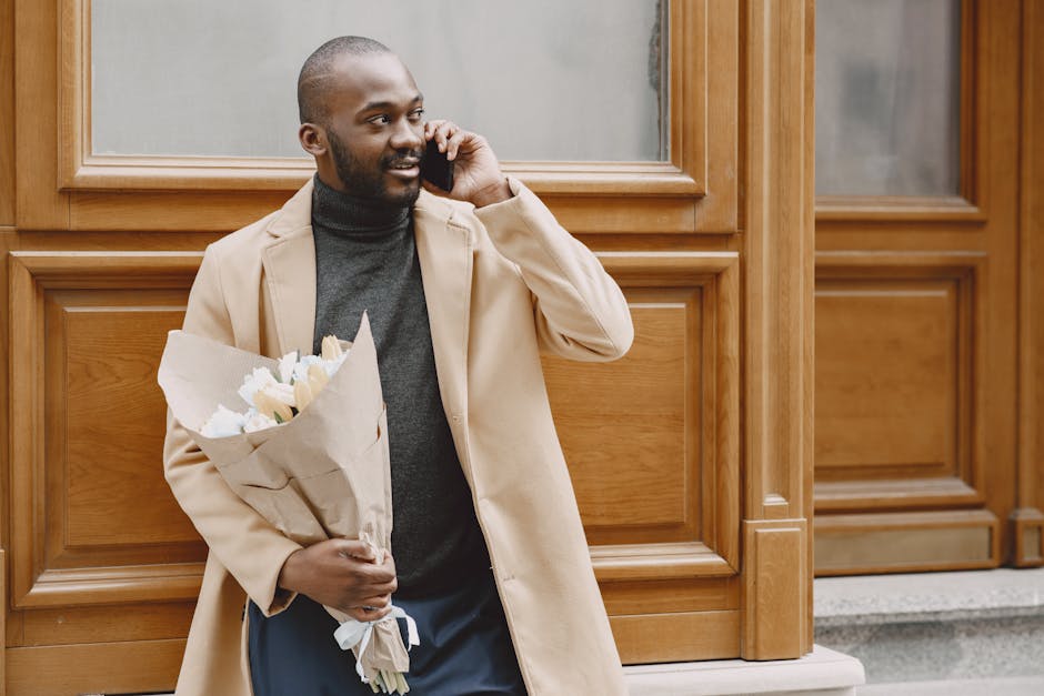 A man with dark skin, short hair, and a beard is standing outdoors against a wooden-clad building, holding a bouquet of fresh white flowers wrapped in plain brown paper. He is wearing a beige overcoat