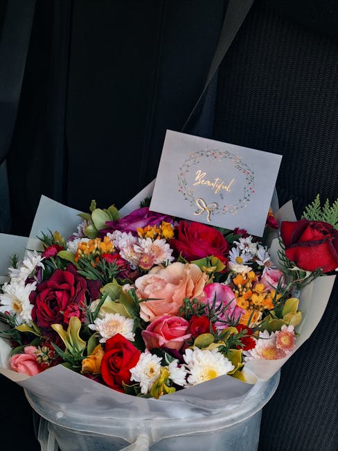 A vibrant bouquet of fresh flowers arranged in a round display, featuring red roses, pink carnations, white daisies, yellow and purple blossoms, and green foliage. The bouquet is wrapped in clear cell