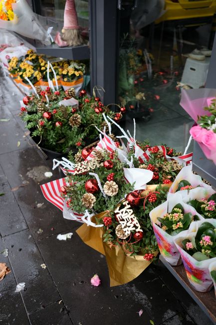 A festive outdoor display of Christmas-themed floral arrangements and bouquets placed on the pavement outside Florist Bowes Park. The arrangements feature a variety of evergreen branches, pinecones, r